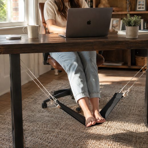 Black fabric foot hammock suspended under a wooden office desk for relaxation while working.