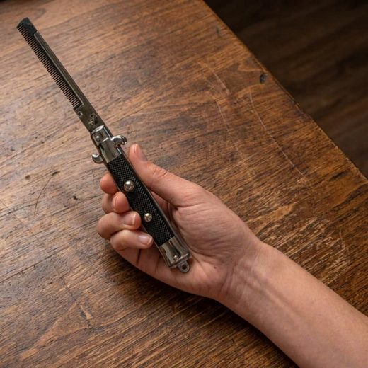 Hand holding a metallic switchblade style comb with a black textured grip on a wooden surface.